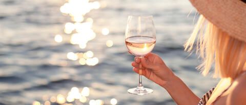 Frau hält Weinglas mit Weisswein in der Hand. Im Hintergrund die Sonne über Wasser.
