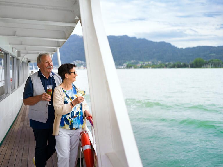 Ein älterer Mann und seine Frau stehen an der Reling eines Schiffs mit Getränken in der Hand. Beide schauen über den See.