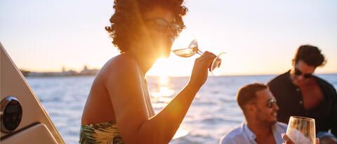 Frau mit einem Glas Wein auf Deck von einem Schiff der BSB. Im Hintergrund sind zwei Männer und der Bodensee.