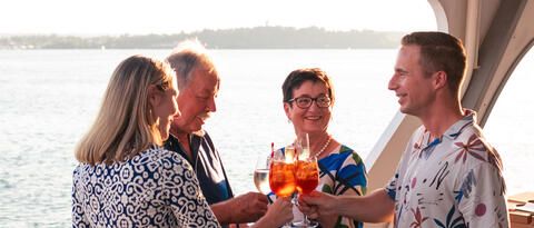 Familie mit vier Personen steht auf einem Schiff der BSB zusammen, alle haben ein Getränk in der Hand, sie stoßen an, die Sonne scheint, im Hintergrund der See.