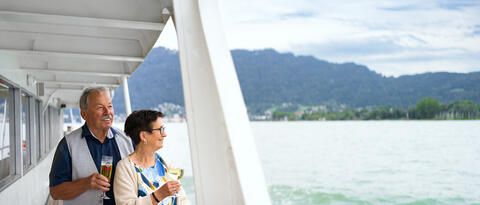 Ein älterer Mann und seine Frau stehen an der Reling eines Schiffs mit Getränken in der Hand. Beide schauen über den See.