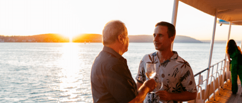 Vater und Sohn stehen an einer Reling, im Hintergrund geht hinter dem See die Sonne unter, beide trinken einen Weisswein.