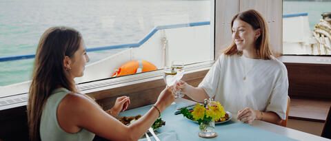 Zwei Frauen bei einer Fahrt mit dem Schiff, die anstoßen und einen Lachsteller essen