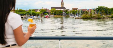 Eine Frau mit Getränk in der Hand blickt vom Schiff aus auf Radolfzell