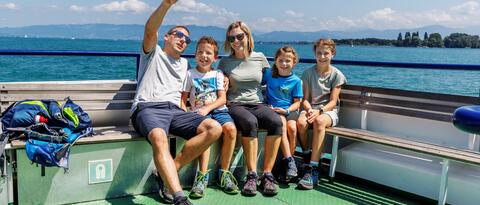 Eine Familie mit 3 Kindern sitzt auf der Bank eines Schiffs, das auf dem blauen Bodensee fährt. Der Vater zeigt mit seinem Finger in den Himmel.