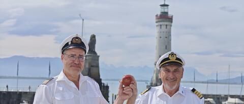 Zwei Kapitäne halten vor Kulisse des Lindauer Hafens gemeinsam einen roten Apfel in die Höhe.