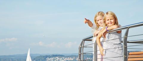 Zwei Kinder an Bord einer Fähre, im Hintergrund das blaue Wasser des Bodensees und ein Segelboot.