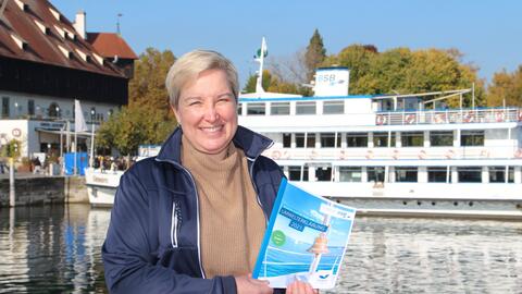 Frau Skoglund von den BSB, steht am Konstanzer Hafen und hält ein Heft von der BSB in der Hand. Im Hintergrund steht ein Schiff von der weißen Flotte.