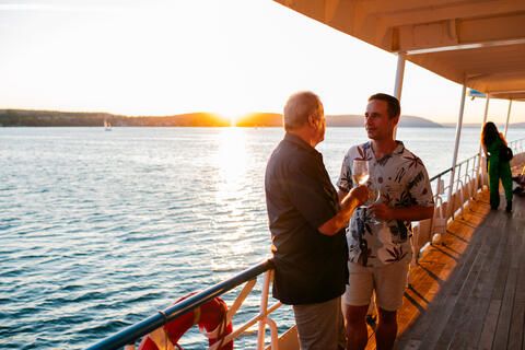 Zwei Männer stehen an der Reling auf einem Schiff der BSB, sie unterhalten sich, im Hintergrund geht hinter dem See die Sonne unter.