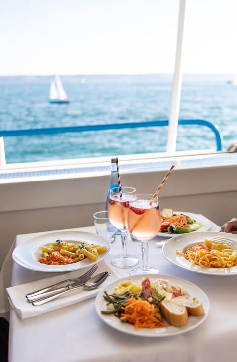 Ein reichlich gedeckter Tisch mit Pasta, Salat und Getränken sowie mit Blick auf den Bodensee