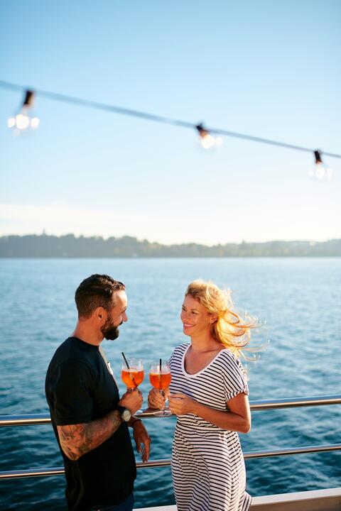 Ein Mann und eine Frau stehen mit Aperol in der Hand an der Reling