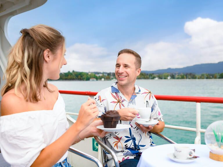 Junges Paar sitzt auf dem Schiff an der Reling auf einem Schiff der BSB, die Frau isst einen Muffin, der Mann trinkt einen Kaffee, beide lachen.