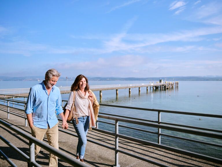 Mann und Frau laufen Hand in Hand auf dem Steg zur Insel Mainau, die Sonne scheint, der Himmel ist blau. Im Hintergrund sieht man den Bodensee.