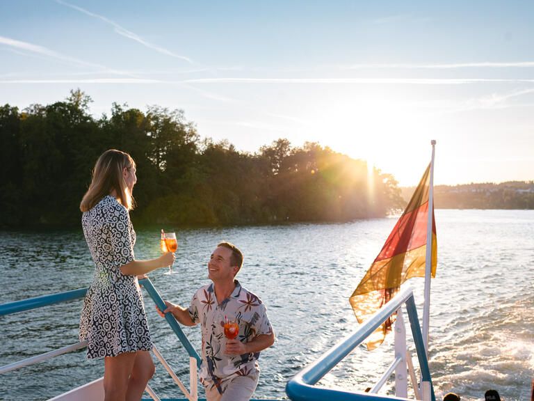Ein Mann und eine Frau im Abendlicht an Bord. In ihrer Hand halten sie ein Getränk.