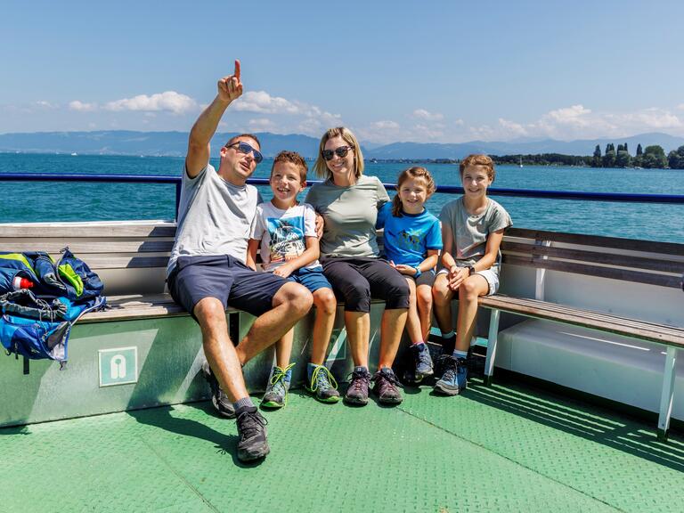 Eine Familie mit 3 Kindern sitzt auf der Bank eines Schiffs, das auf dem blauen Bodensee fährt. Der Vater zeigt mit seinem Finger in den Himmel.