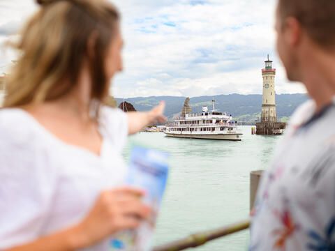 Junges Paar am Hafen in Lindau und schaut auf die Hafeneinfahrt, wo ein Schiff einfährt. Die Frau zeigt auf das Schiff.