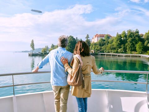 Mann und Frau stehen an der Reling eines BSB Schiffs und schauen über den See auf die Insel Mainau bei blauem Himmel. Am Himmel sieht man einen Zeppelin.
